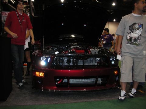 1998 Ford Crown Victoria - dark red Ford Mustang with its hood up and headlights on sits between two men on a green-carpeted show floor.