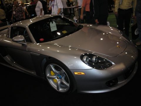 1998 Ford Crown Victoria - silver Porsche Carrera GT supercar with yellow brake calipers sits under spotlights amid spectators at an indoor show.