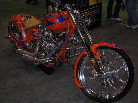 1998 Ford Crown Victoria - custom chopper motorcycle painted orange with blue flames sits on chrome wheels at an indoor show display.