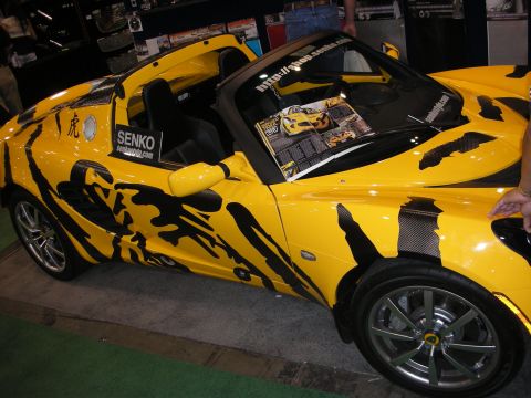 1998 Ford Crown Victoria - yellow Lotus Elise roadster with black tribal graphics and a SENKO decal sits on display with an info sheet on the windshield.
