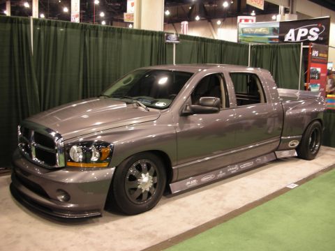1998 Ford Crown Victoria - lowered gray Dodge Ram crew-cab pickup with black wheels sits on a green-carpeted booth in front of an APS banner at a show.