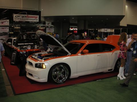 1998 Ford Crown Victoria - white Dodge Charger with orange accent stripes, chrome wheels and open hood is displayed beside a model at an indoor car show.