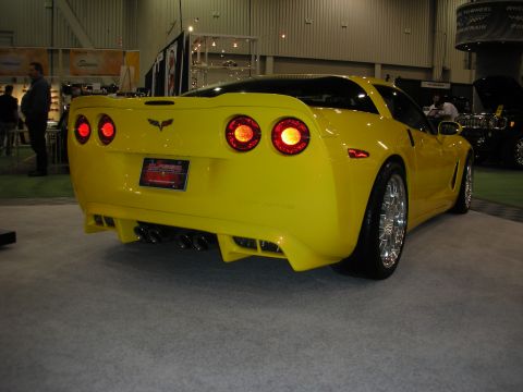 1998 Ford Crown Victoria - Rear three-quarter view of a yellow Chevrolet Corvette coupe with round taillights, quad exhaust and chrome wheels on a show floor.