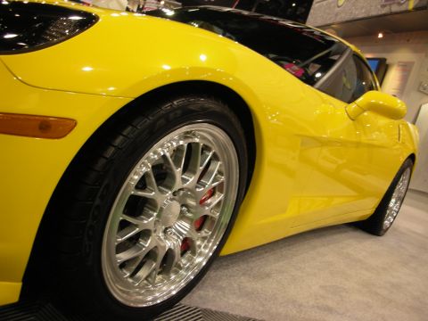 1998 Ford Crown Victoria - Detail of a yellow Corvette's front fender and polished mesh-style wheel with a red brake caliper at an indoor display.