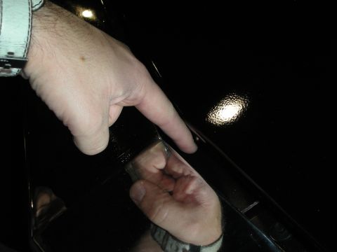 1998 Ford Crown Victoria - person's hand points at a dark, glossy vehicle surface reflecting a small light in a dimly lit setting.
