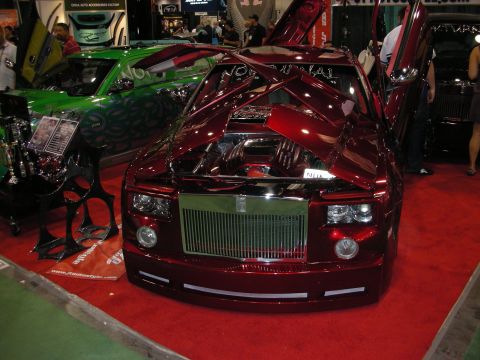 1998 Ford Crown Victoria - candy-red custom Rolls-Royce Phantom with raised hood, chrome grille and big chrome wheels sits beside a green car at a show.
