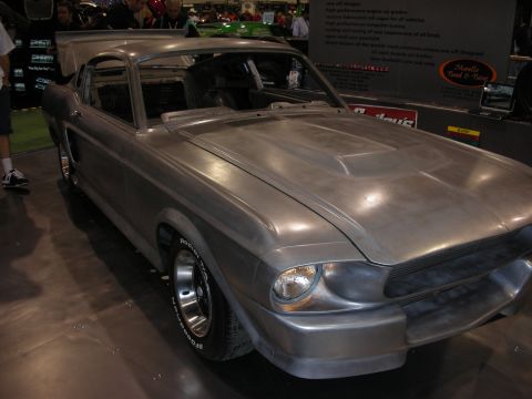 1998 Ford Crown Victoria - unpainted bare-metal fastback muscle car with mag wheels displayed at an indoor automotive trade show booth.
