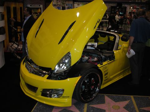 1998 Ford Crown Victoria - bright yellow custom convertible with its hood up and a scissor door raised, on display over a walk-of-fame star floor.