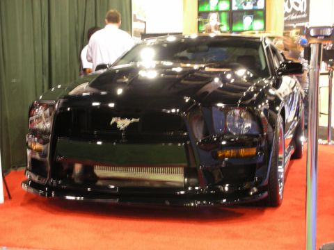 1998 Ford Crown Victoria - black custom Ford Mustang shown front-on with running-pony badge, large intercooler and aggressive fascia on a red display floor.