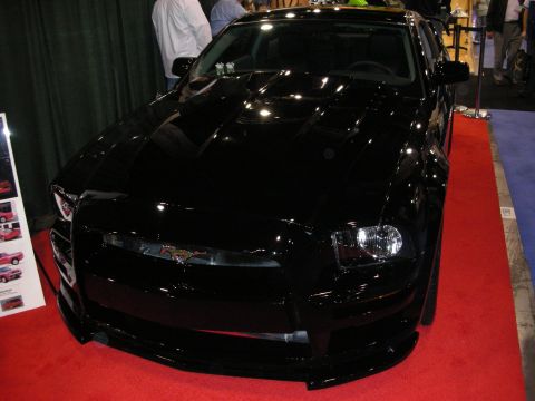 1998 Ford Crown Victoria - glossy black custom Ford Mustang with running-pony badge displayed on a red carpet at an indoor automotive show.
