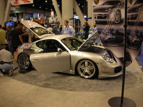 1998 Ford Crown Victoria - silver Porsche sports coupe with doors and hood open displayed beside a Cayman banner at a crowded indoor trade show.