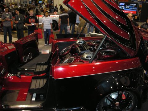 1998 Ford Crown Victoria - Candy-red custom show car with its hood raised to reveal a ringed speaker array, displayed amid a crowd at a trade show.