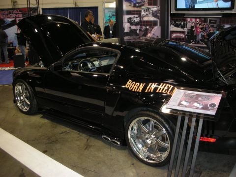 1998 Ford Crown Victoria - black custom coupe shown side-on with chrome wheels and a gold 'BORN IN HELL' decal on the rear quarter at an indoor show.
