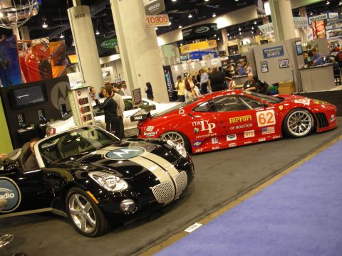1998 Ford Crown Victoria - red Ferrari 'of Houston' #62 race car displayed beside a black-and-silver striped roadster at an indoor automotive show.