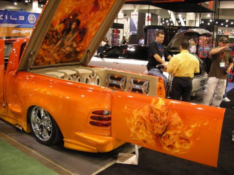1998 Ford Crown Victoria - orange flame-airbrushed lowered pickup with raised bed cover revealing a chrome speaker install at a car-audio trade show.