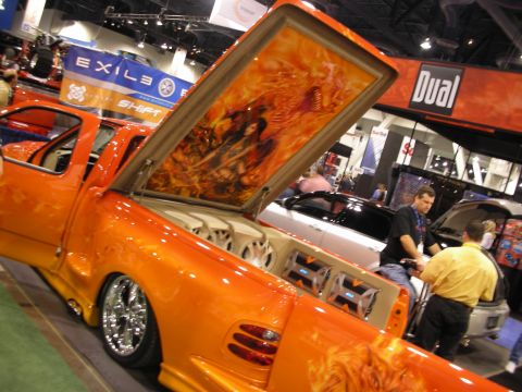 1998 Ford Crown Victoria - Orange flame-painted lowered pickup with an airbrushed fantasy mural on its raised bed cover at a car-audio show booth.