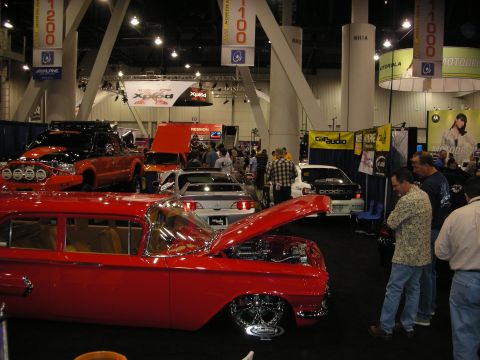 1998 Ford Crown Victoria - red classic car parked on a carpeted convention floor with raised-hood trucks and crowds at a car audio trade show.