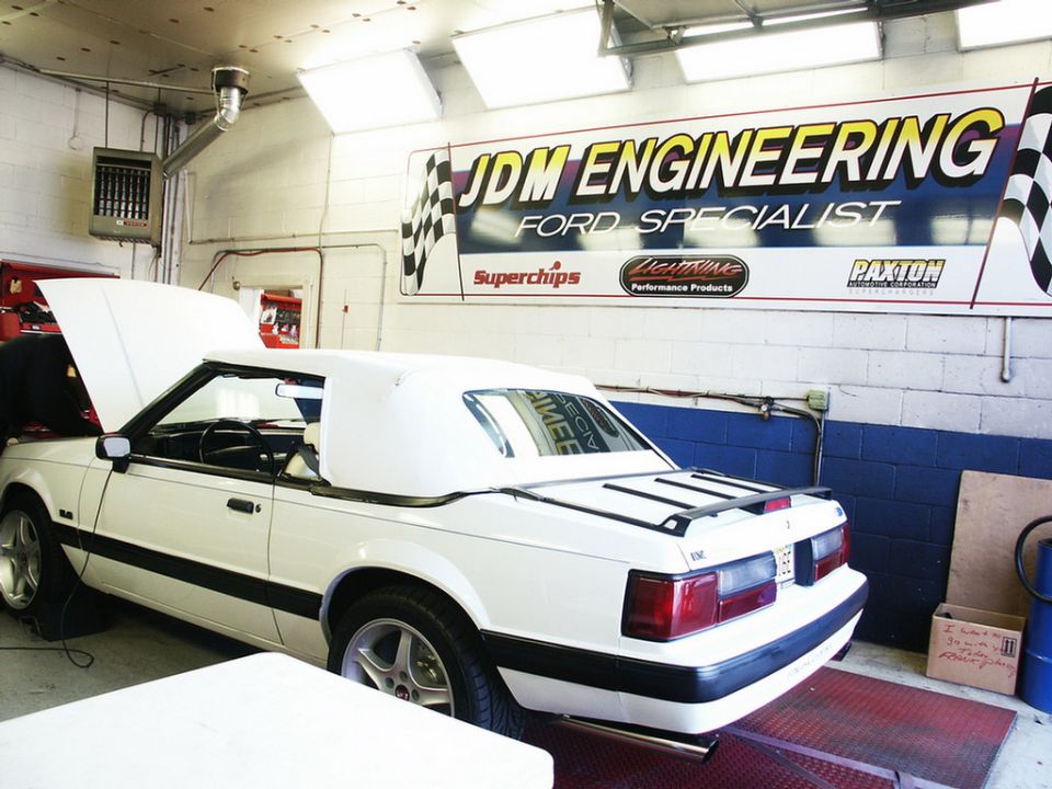 White convertible with the hood up, parked on a dynamometer in a garage. The car has a black trim and a black spoiler