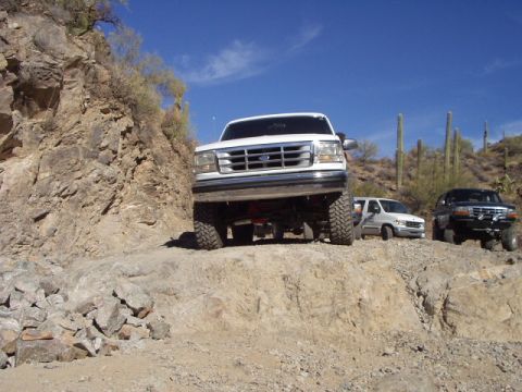 1993 Ford Bronco - A white Ford Bronco navigates a challenging off-road path, with other vehicles visible in the distance.