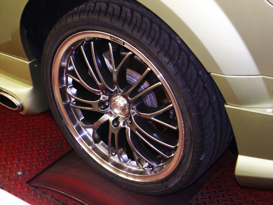 Close-up of a shiny custom wheel with a low-profile tire on a gold car. The car has a side skirt and exhaust tip visible