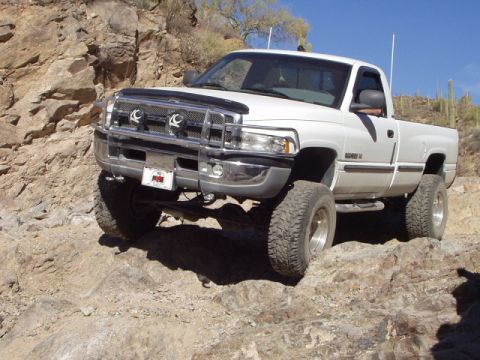 1993 Ford Bronco - White Dodge Ram pickup truck navigates a rocky off-road trail under a clear blue sky.