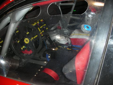 1998 Ford Crown Victoria - bare Ferrari race-car interior viewed through glass, showing a roll cage, flat-bottom wheel with prancing-horse badge and red harness.