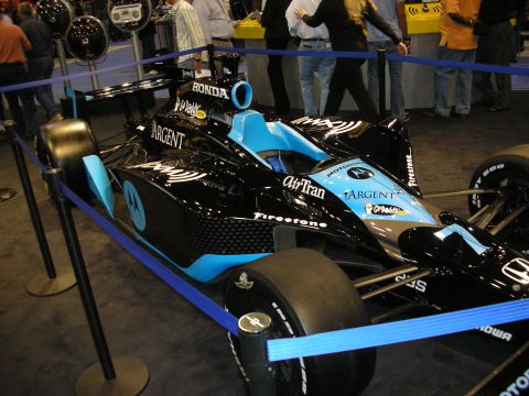 1998 Ford Crown Victoria - black-and-blue Honda-powered IndyCar on Firestone tires, displayed behind blue rope barriers with AirTran and Argent decals.