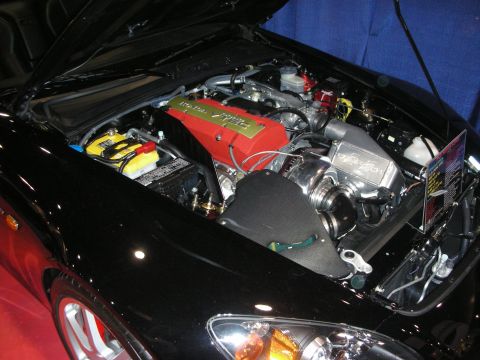 1998 Ford Crown Victoria - open engine bay of a black sports car shows a red plug-cover, polished metal piping and a yellow battery at an indoor show.