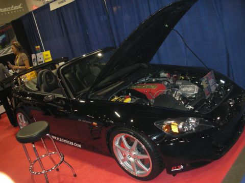 1998 Ford Crown Victoria - black two-seat convertible sits with its hood raised on red carpet at an indoor auto show, a PLX Devices stool beside it.