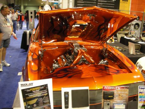 1998 Ford Crown Victoria - glossy orange custom car with airbrushed flames has its trunk raised over a chrome engine, shown above audio displays at a show.