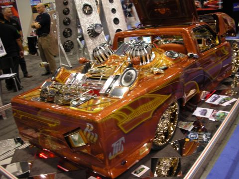 1998 Ford Crown Victoria - candy-orange custom lowrider truck with engraved chrome, gold spoke wheels and graphics sits on mirrored flooring at a show.