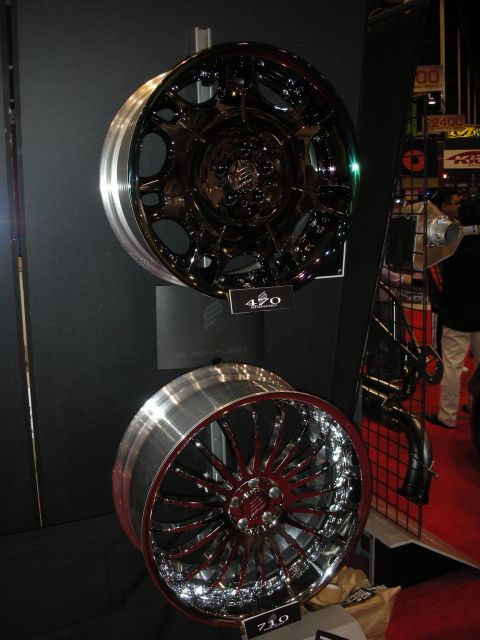 1998 Ford Crown Victoria - Two custom wheels on a vendor stand: a dark mesh wheel above and a chrome multi-spoke wheel with red accents below.