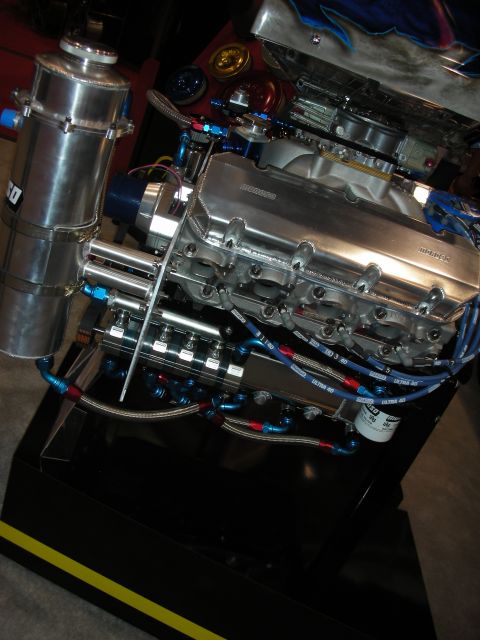 1998 Ford Crown Victoria - polished V8 crate engine with a carburetor and blue braided fittings sits on a black display base at a show.