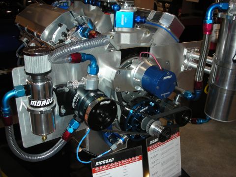 1998 Ford Crown Victoria - Moroso display engine assembly with dry-sump pumps, blue and red braided lines and an aluminum reservoir on a booth stand.