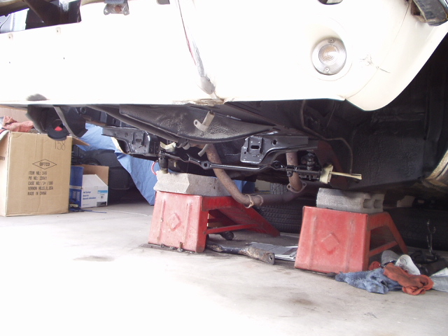 Underside of a vehicle on jack stands, showing suspension and exhaust components. Cardboard boxes are nearby