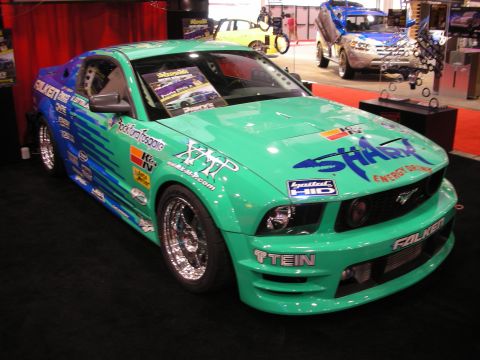 1998 Ford Crown Victoria - teal-and-blue widebody Ford Mustang covered in sponsor decals sits on display at an indoor automotive trade show.