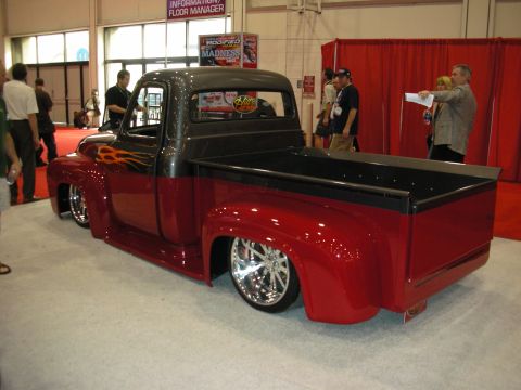 1998 Ford Crown Victoria - lowered dark-red classic pickup truck with flame graphics sits on a carpeted trade-show floor as people walk by.