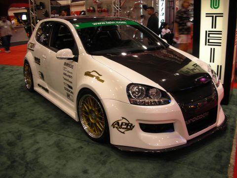 1998 Ford Crown Victoria - white-and-carbon widebody Volkswagen Golf hatchback with gold wheels and sponsor decals at an indoor trade show.