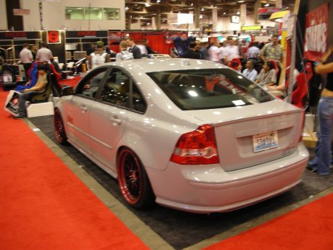 1998 Ford Crown Victoria - lowered silver Volvo sedan with red wheels sits rear-three-quarter on a busy indoor show floor.