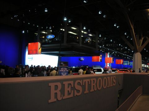 1998 Ford Crown Victoria - dimly lit convention hall shows a Ford booth above a wall marked RESTROOMS with a crowd below.
