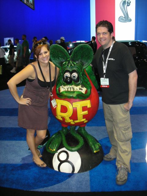 1998 Ford Crown Victoria - woman and a man pose on either side of a large green Rat Fink statue mounted on an 8-ball at a trade show.