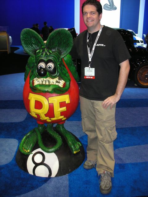 1998 Ford Crown Victoria - man stands smiling beside a large green Rat Fink character statue mounted on a black 8-ball at an indoor show.