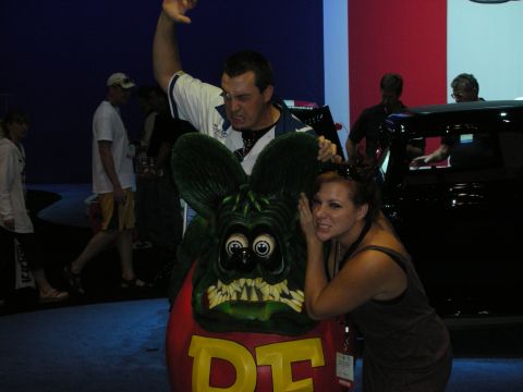 1998 Ford Crown Victoria - man raises his fist and a woman leans in as they pose behind a large green Rat Fink statue on a show floor.
