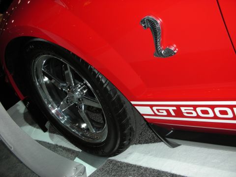 1998 Ford Crown Victoria - Close-up of a red GT500's rear quarter panel showing a chrome five-spoke wheel, Cobra snake emblem and white GT 500 racing stripe.