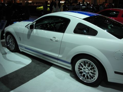 1998 Ford Crown Victoria - Side profile of a white sports coupe with blue over-the-top racing stripes and chrome mesh wheels at an indoor auto show.