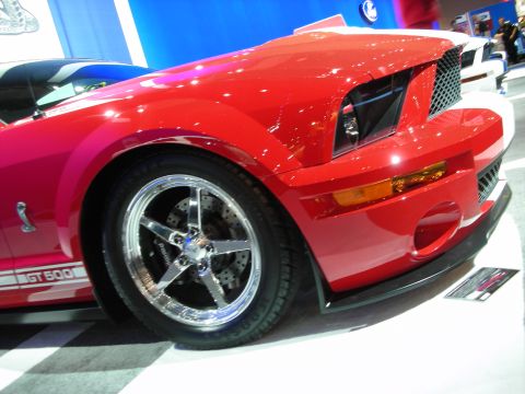 1998 Ford Crown Victoria - Low front three-quarter view of a red GT500 with chrome five-spoke wheel and GT 500 rocker stripe on a checkered show floor.