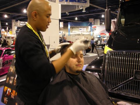 1998 Ford Crown Victoria - man wearing latex gloves and a lanyard styles a seated man's hair beside a black truck grille at an indoor trade show.