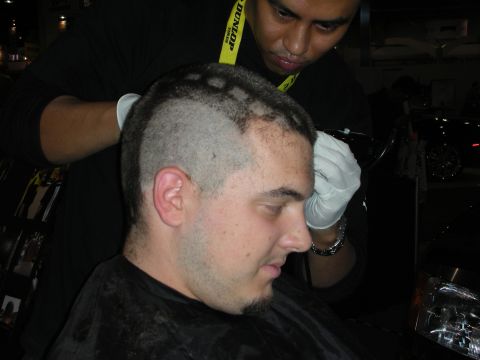 1998 Ford Crown Victoria - gloved barber in a Dunlop lanyard clips a pattern into the side of a seated young man's closely cropped hair.