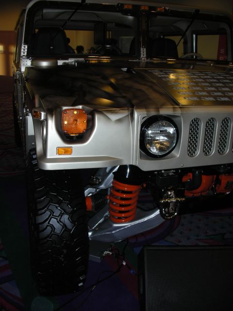 1998 Ford Crown Victoria - Front corner of a custom silver Hummer H1 showing the round headlight, slatted hood, amber turn signal and an exposed orange coil-over.