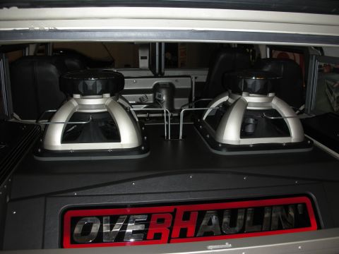 1998 Ford Crown Victoria - Two large subwoofers in chrome and black enclosures mounted in a vehicle's rear cargo area above a red OVERHAULIN' nameplate.